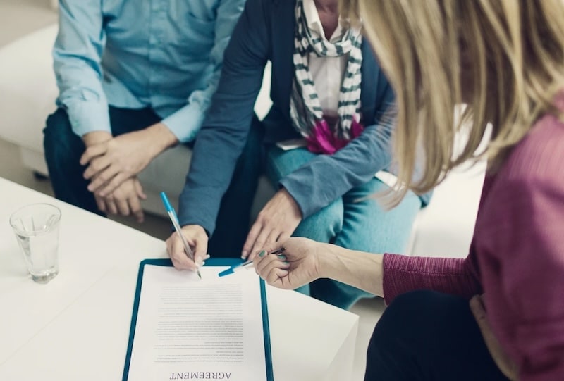 A person signing a legal agreement document on a clipboard while sitting with two other professionals during a business meeting or consultation.