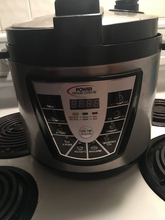 A stainless steel Power Pressure Cooker XL sitting on a white stovetop, showing the digital control panel with various cooking presets like slow cook, rice, and beans.