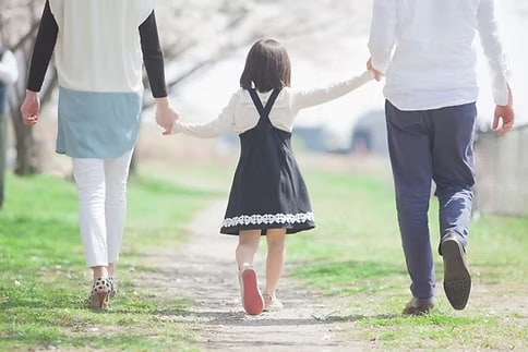 Rear view of a young girl in a black pinafore dress holding hands with her parents as they walk together along a sunlit path in a park.