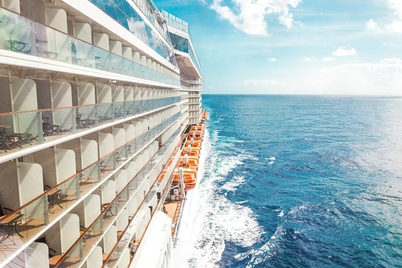 Side view of a large modern cruise ship sailing on the bright blue ocean, showcasing rows of private balconies and orange lifeboats under a sunny sky.