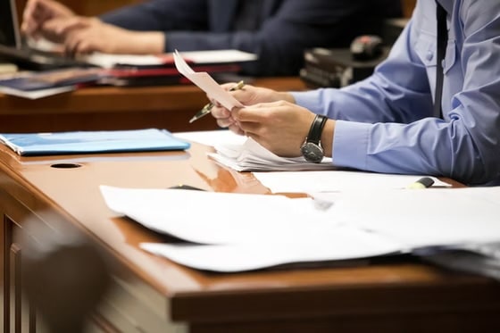 Close-up of a person in a blue uniform shirt reviewing legal documents and papers at a wooden desk in a courtroom or official setting.