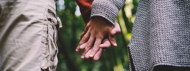 Close-up of a couple holding hands while walking through a forest, with one person wearing cargo pants and the other wearing a grey knit sweater.