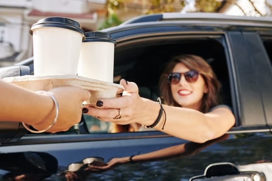 A woman in sunglasses reaching out from a car window to receive two takeaway coffees in a cardboard carrier.