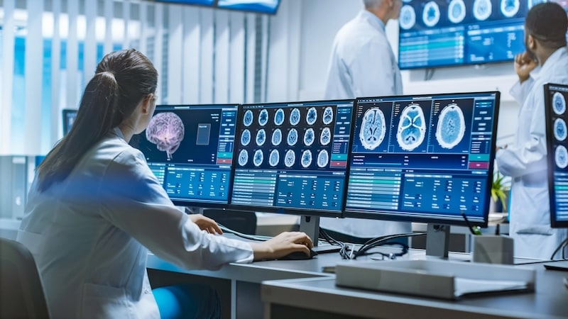 A female scientist in a white lab coat analyzing brain scan results and neurological data on multiple computer monitors in a high-tech medical research facility.