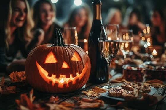 A glowing Jack-o'-lantern on a wooden table set for a Halloween party, featuring a wine bottle, glasses of champagne, and snacks with a blurred group of people laughing in the background.