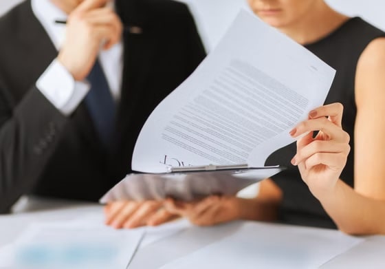 Two business professionals in formal attire reviewing a legal contract on a clipboard during a meeting.