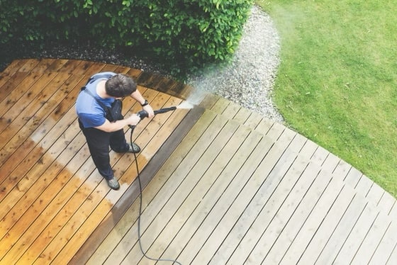 High-angle view of a person using a pressure washer to clean a curved wooden deck, showing the contrast between the dirty grey wood and the clean brown surface.