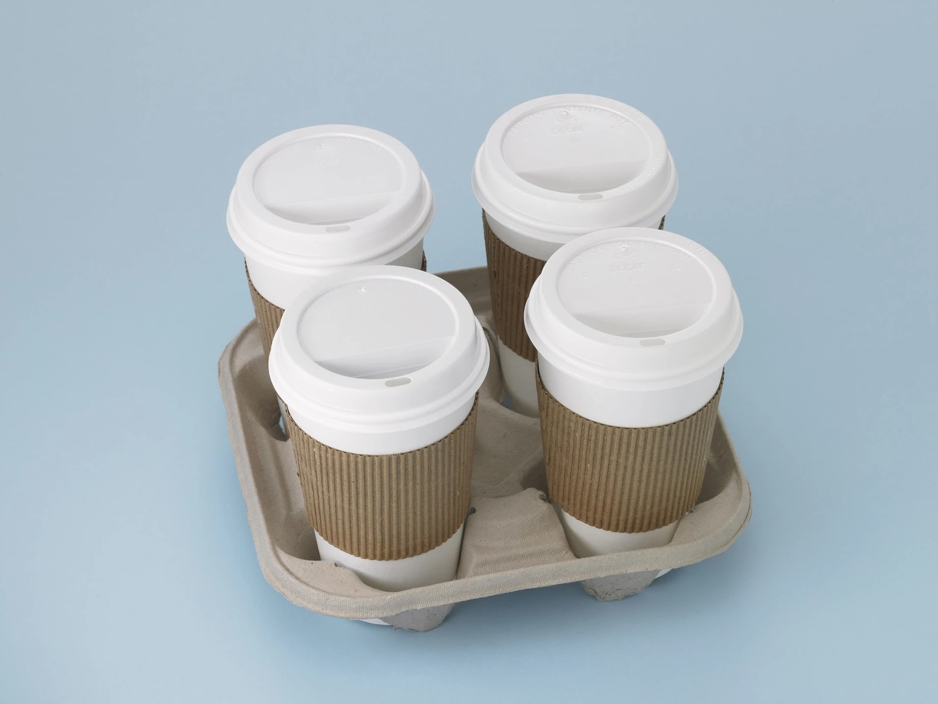 Four disposable coffee cups with white lids and brown cardboard sleeves in a four-cup carrier against a light blue background.