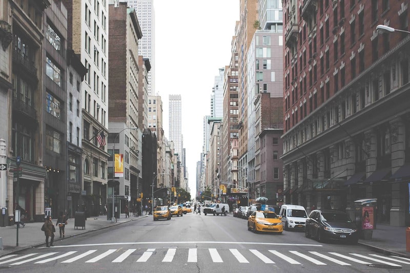 A street-level, perspective view looking down a busy New York City thoroughfare, featuring iconic yellow taxis, parked cars, and pedestrians crossing a white-striped crosswalk flanked by tall urban buildings.