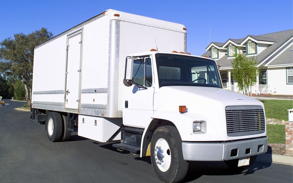 A three-quarter front view of a white box truck parked on a residential street. The vehicle features a large, windowless cargo area with a side access door, parked in front of a suburban home under a clear blue sky.