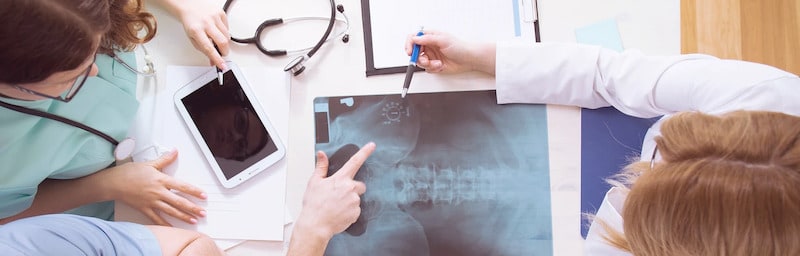Top-down view of three medical professionals in scrubs and white coats gathered around a desk, collaborating on a patient's case while reviewing a spinal X-ray and a digital tablet.