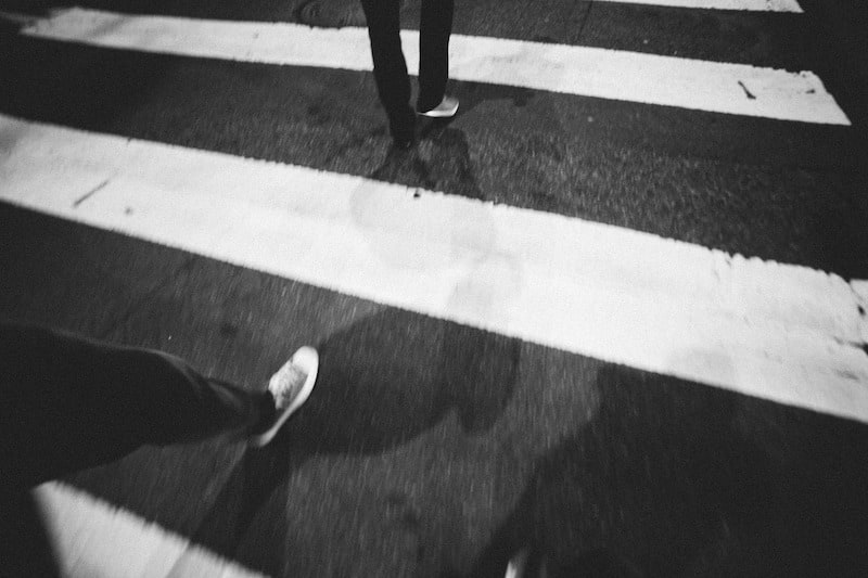 A high-contrast, black-and-white street photography shot taken from a low-angle perspective. The image features the legs and feet of pedestrians crossing a white-striped crosswalk at night. The dark shadows cast against the asphalt and the grainy texture of the film-style aesthetic create a moody, urban atmosphere.