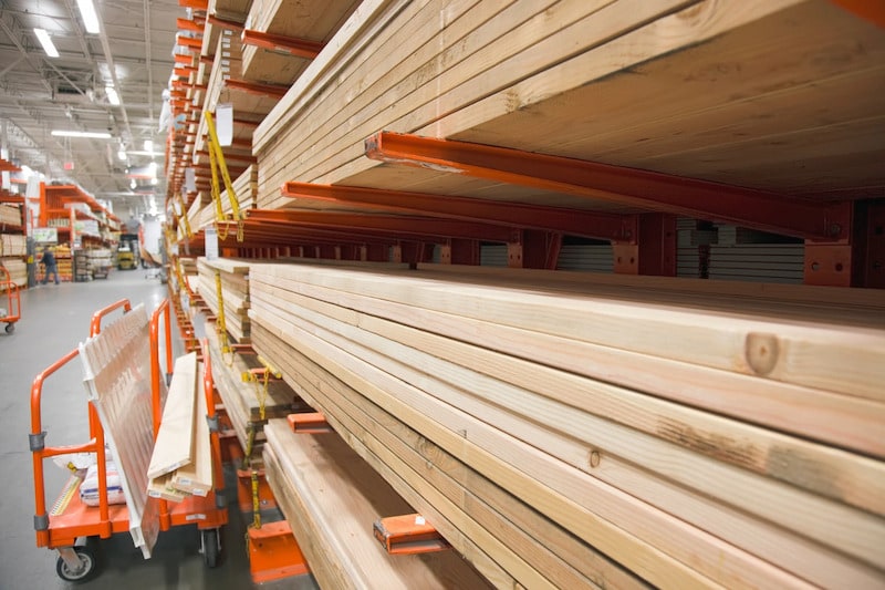 A high-angle, perspective shot of stacks of lumber organized on orange industrial cantilever racks in a home improvement store aisle, with a loaded orange cart visible in the foreground.
