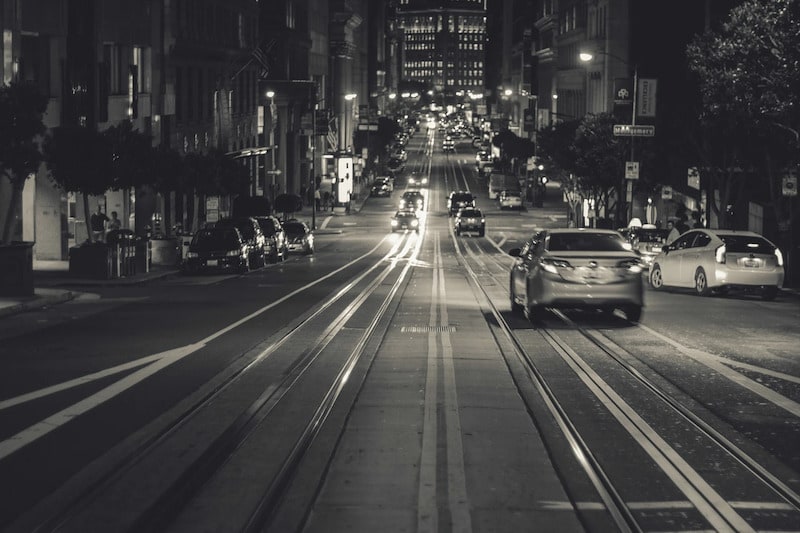 Black and white night photography of a busy San Francisco street showing cable car tracks leading into the distance with cars driving under glowing city lights.