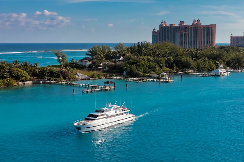 Aerial view of a large white yacht cruising through turquoise tropical waters toward a lush island with docks and the Atlantis Bahamas resort in the background.