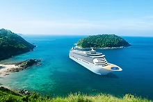 Aerial view of a large white cruise ship sailing through turquoise waters near lush tropical islands under a clear blue sky.