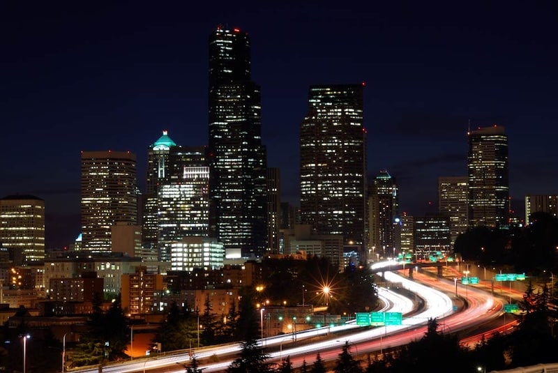 A night-time city skyline featuring illuminated skyscrapers, including the Columbia Center in Seattle, with a long-exposure shot of a highway in the foreground showing light trails from passing vehicles.