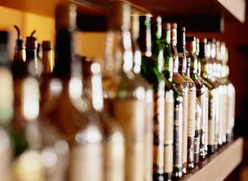 A shallow-depth-of-field shot of various liquor bottles lined up on a wooden bar shelf, with the focus on the labels of the bottles in the mid-ground while the foreground remains blurred.