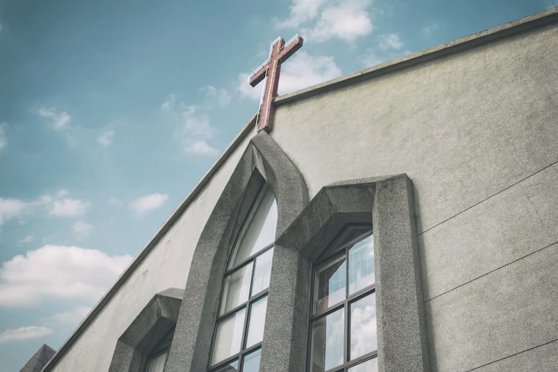 A low-angle, perspective shot of a modern church building with a light gray stone or concrete facade. The architecture features prominent pointed gothic-style window frames and is topped with a simple red cross at the peak of the roof. The building is set against a pale blue sky with soft, wispy white clouds.