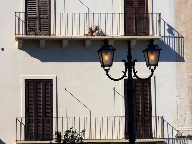 A street-level view of a light-colored building with dark brown shuttered doors and wrought-iron balconies, featuring a classic double-headed black lamppost with glowing lights in the foreground.
