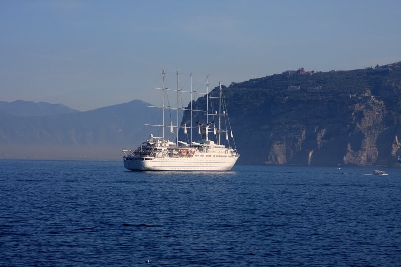 A wide, profile view of a large white five-masted sailing cruise ship (the Wind Surf) gliding across deep blue water. The ship is framed by a backdrop of misty mountains and a steep, rugged cliffside under a clear sky.