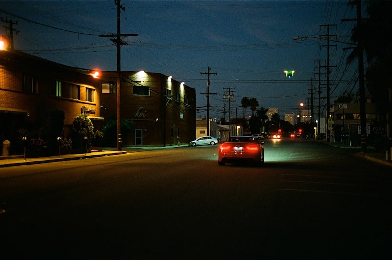 Nighttime street scene in an industrial area featuring a red car with glowing taillights driving past a brick building labeled