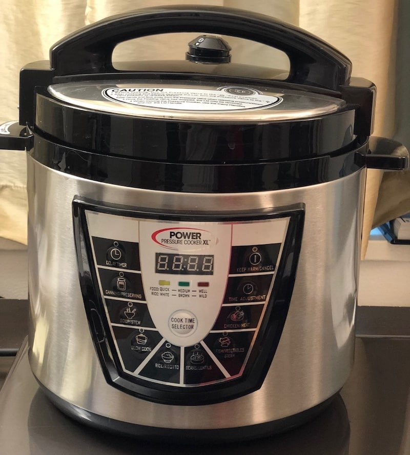 Front view of a stainless steel Power Pressure Cooker XL with its black lid closed, showing the digital interface with buttons for soup, canning, slow cook, and other presets.