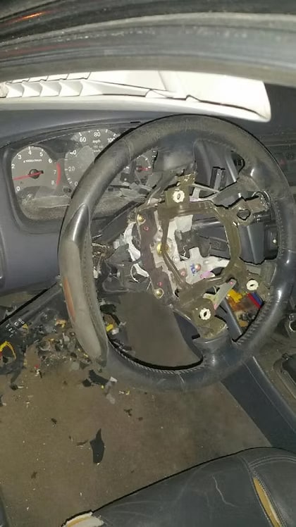 Interior of a vehicle showing a steering wheel with a missing center cover and a deployed or removed airbag assembly, revealing the internal metal frame and wiring.
